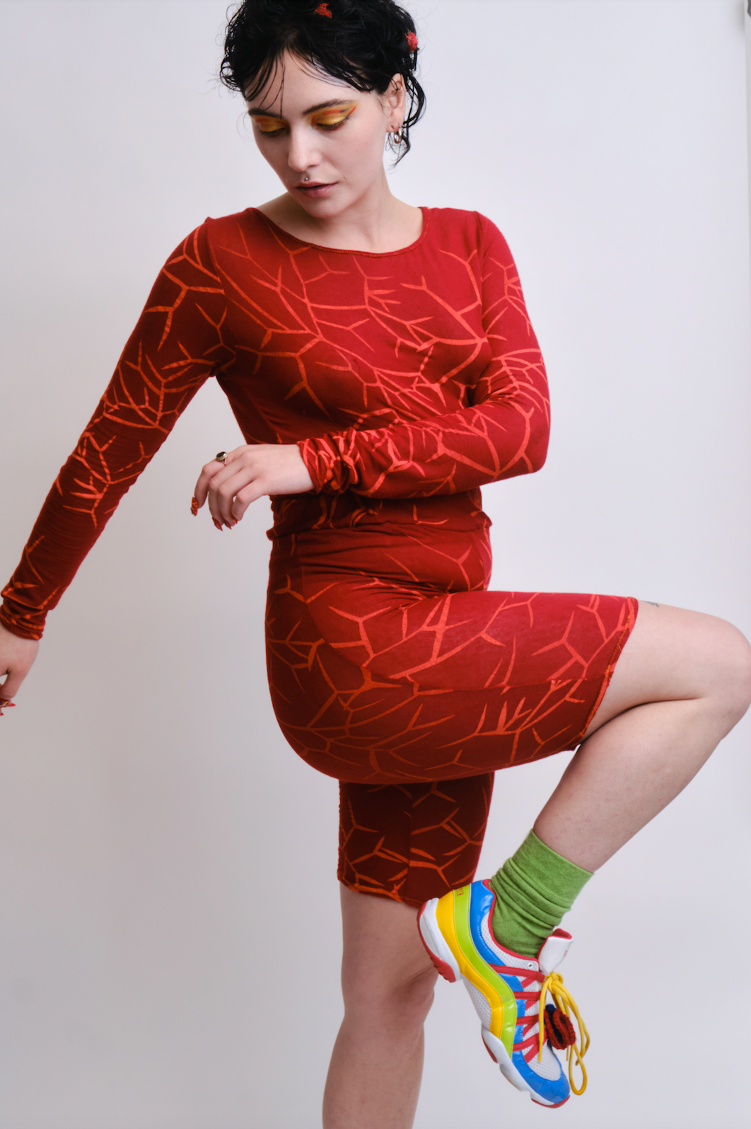 Model wearing a red, abstract printed two-piece active wear set. The model is posed with one arm crossed against their torso, and their opposite leg bent in an upright position. The model wears a long-sleeve shirt with matching biker shorts, green socks and colorful sneakers.