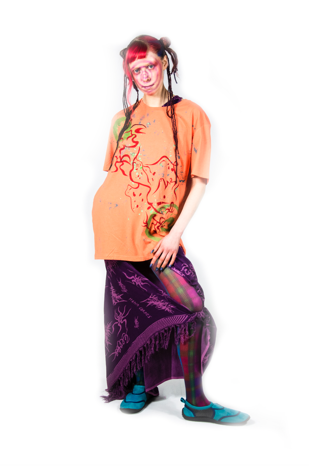 Model is standing with one leg bent while wearing an orange t-shirt and a long purple printed skirt. The orange shirt has abstract red lines on it and the bottom is a long skirt that has a slit near the knee. The model has colorful orange, pink and purple hair pulled back into a sleek bun with strands flowing out. The print of the dark purple outfit contains light, vibrant purplish pink squiggles and drawings that resemble bugs with long legs and leafy plants. 