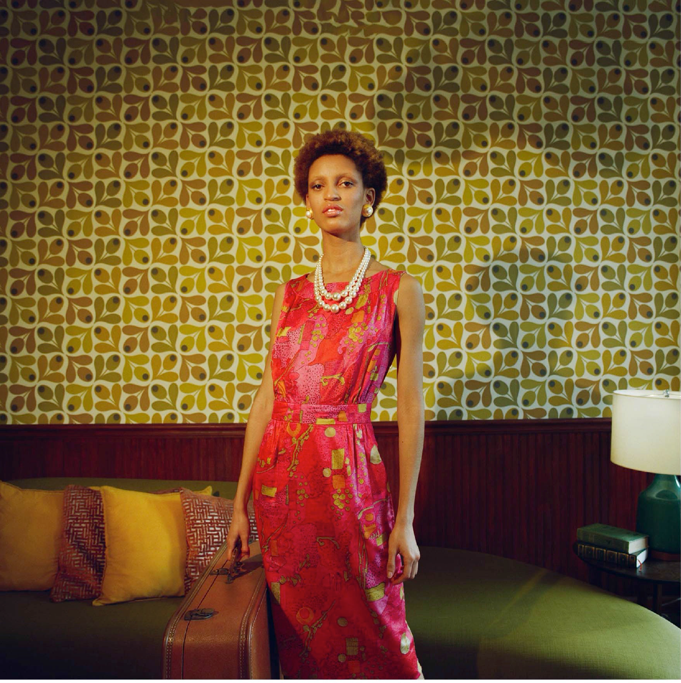 This image shows a woman with a curly pixie haircut, gold stud earrings, a white pearl necklace, and a pinkish red patterned dress. The model is also holding a brown suitcase. The background of the image is a green patterned wallpaper, and a wall couch with green cushions, yellow and orange pillows, and a side table with a lamp.