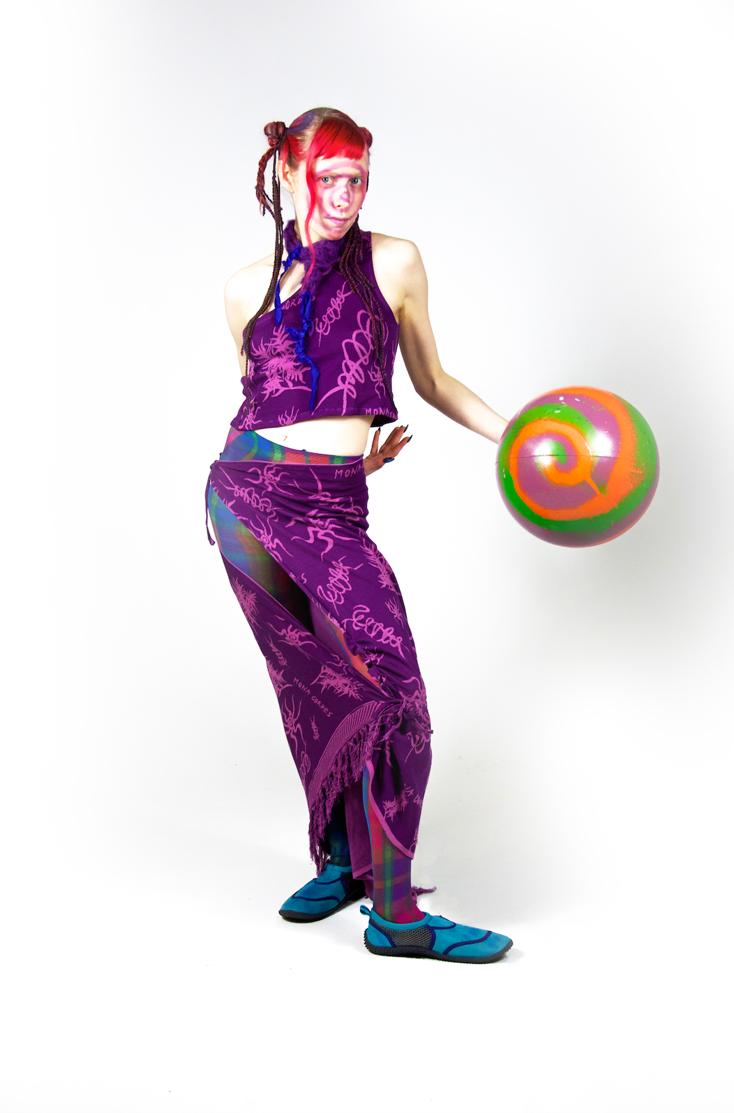 Model is standing with one leg bent and wearing a purple, printed two piece outfit. The top is a cropped tank top, and the bottom is a long skirt that hits right above the shoes. The model has colorful orange, pink and purple hair pulled back into a sleek bun with strands flowing out. The print of the dark purple outfit contains light, vibrant purplish pink squiggles and drawings that resemble bugs with long legs and leafy plants.