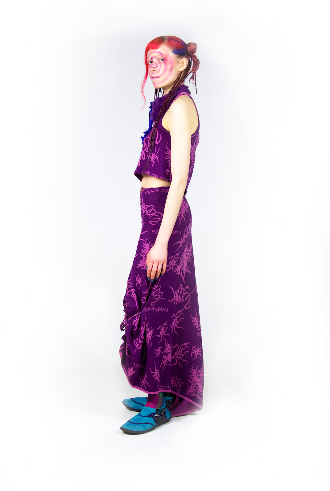 Model is standing sideways wearing a purple, printed two piece outfit. The top is a cropped tank top, and the bottom is a long skirt that hits right above the shoes. The model has colorful orange, pink and purple hair pulled back into a sleek bun with strands flowing out. The print of the dark purple outfit contains light, vibrant purplish pink squiggles and drawings that resemble bugs with long legs and leafy plants. 