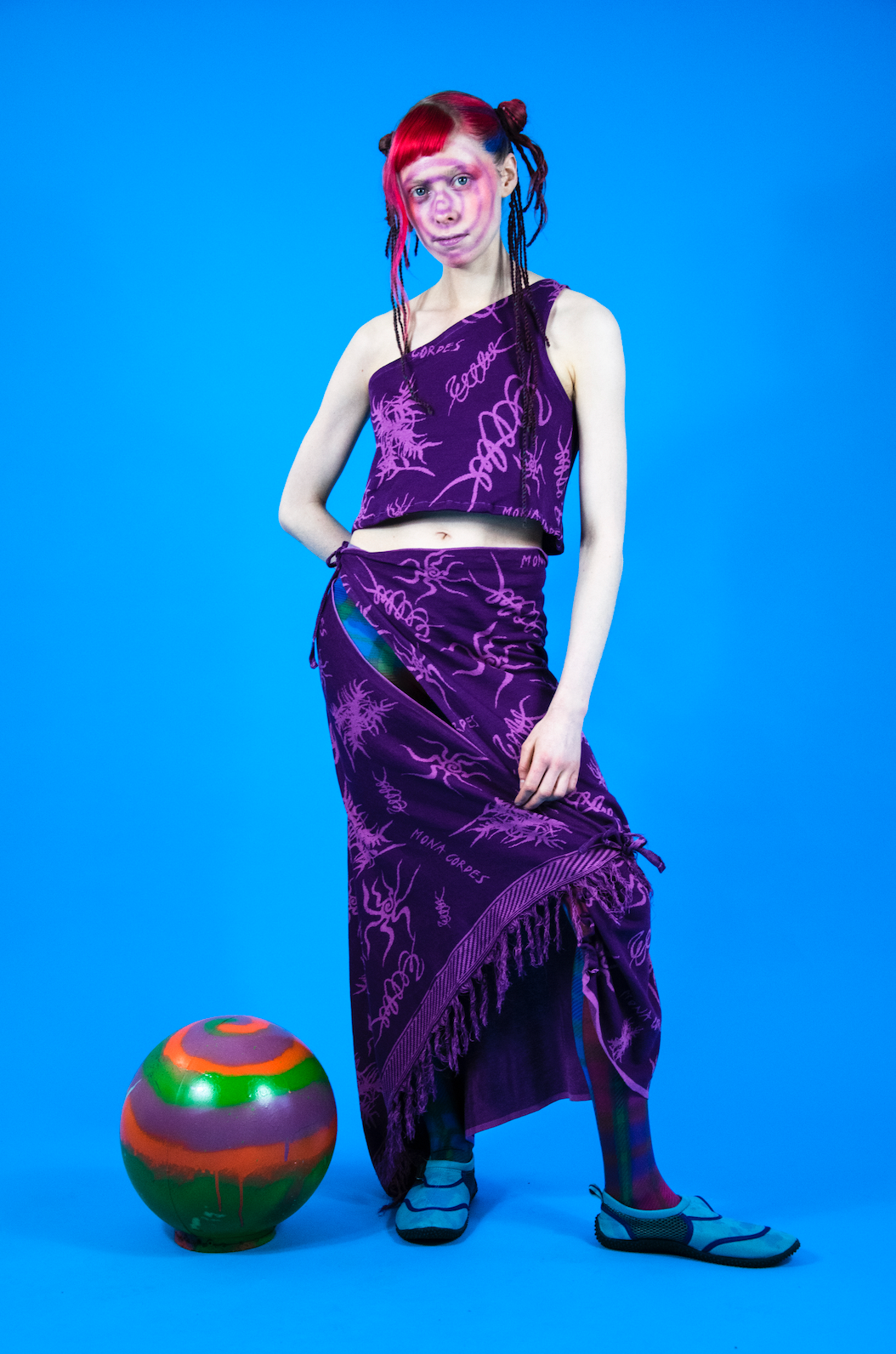 Model is standing sideways wearing a purple, printed two piece outfit. The top is a cropped tank top, and the bottom is a long skirt that hits right above the shoes. The model has colorful orange, pink and purple hair pulled back into a sleek bun with strands flowing out. The print of the dark purple outfit contains light, vibrant purplish pink squiggles and drawings that resemble bugs with long legs and leafy plants.